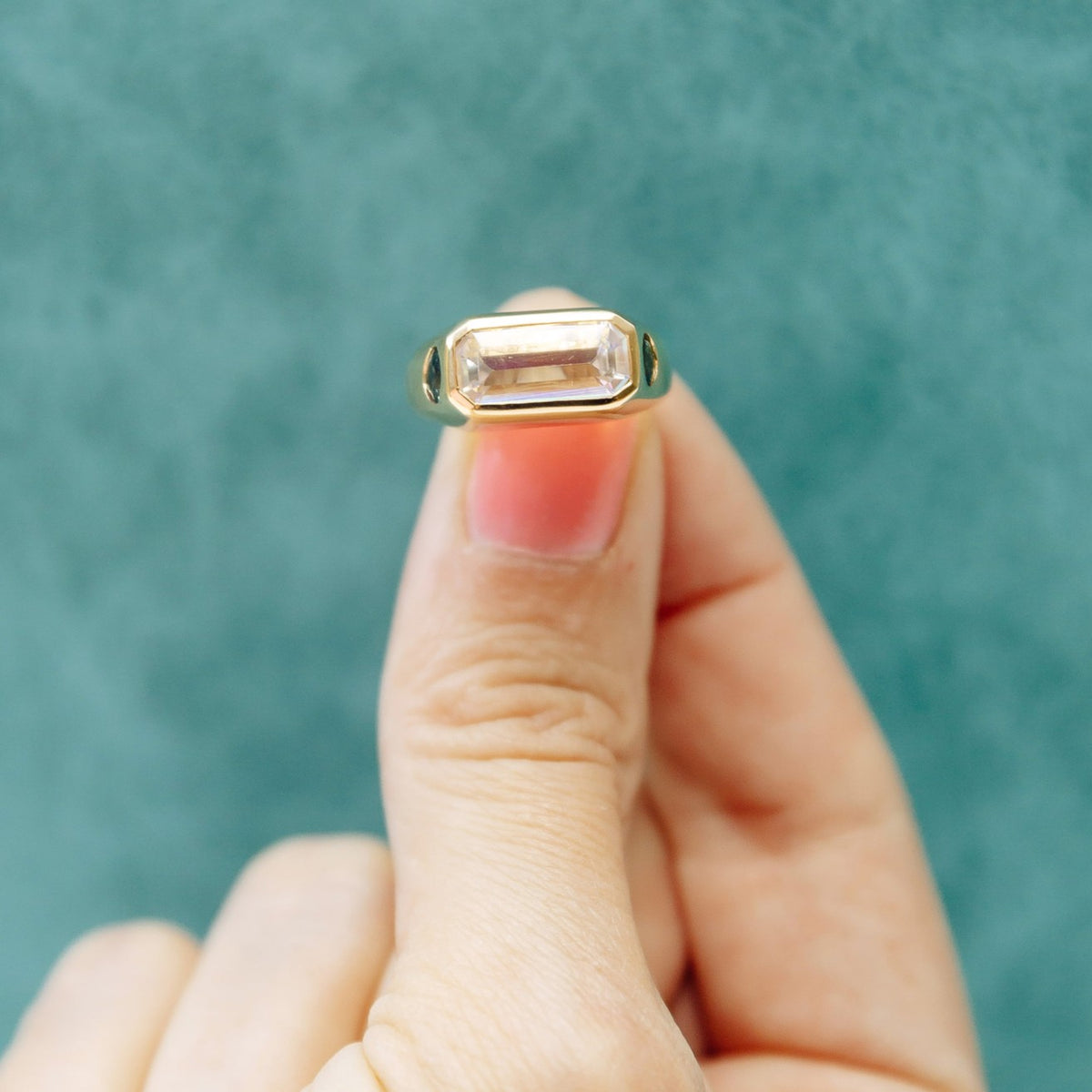 A gold ring with a rectangular clear gemstone held between fingers against a textured teal background.