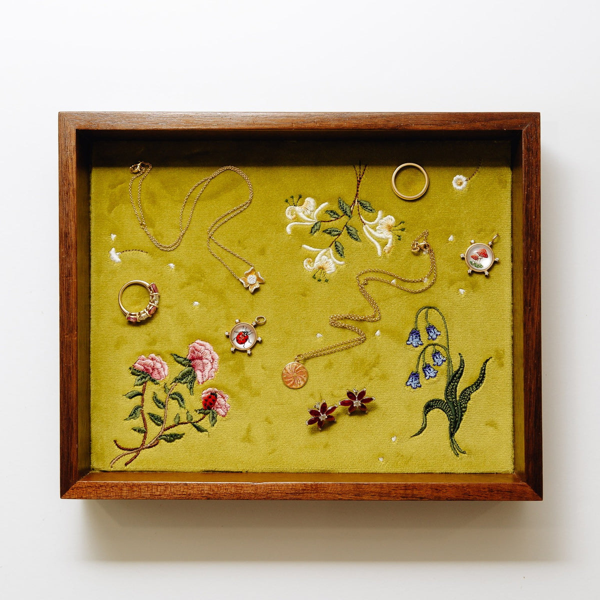 A jewelry tray showcasing various handmade jewelry pieces, including necklaces, rings, and earrings, arranged on a textured olive green velvet background with embroidered floral motifs.
