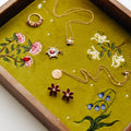 A styled flat-lay of assorted gold jewelry, including necklaces, earrings, and a ring, arranged on an olive green embroidered fabric insert inside a wooden jewelry tray.