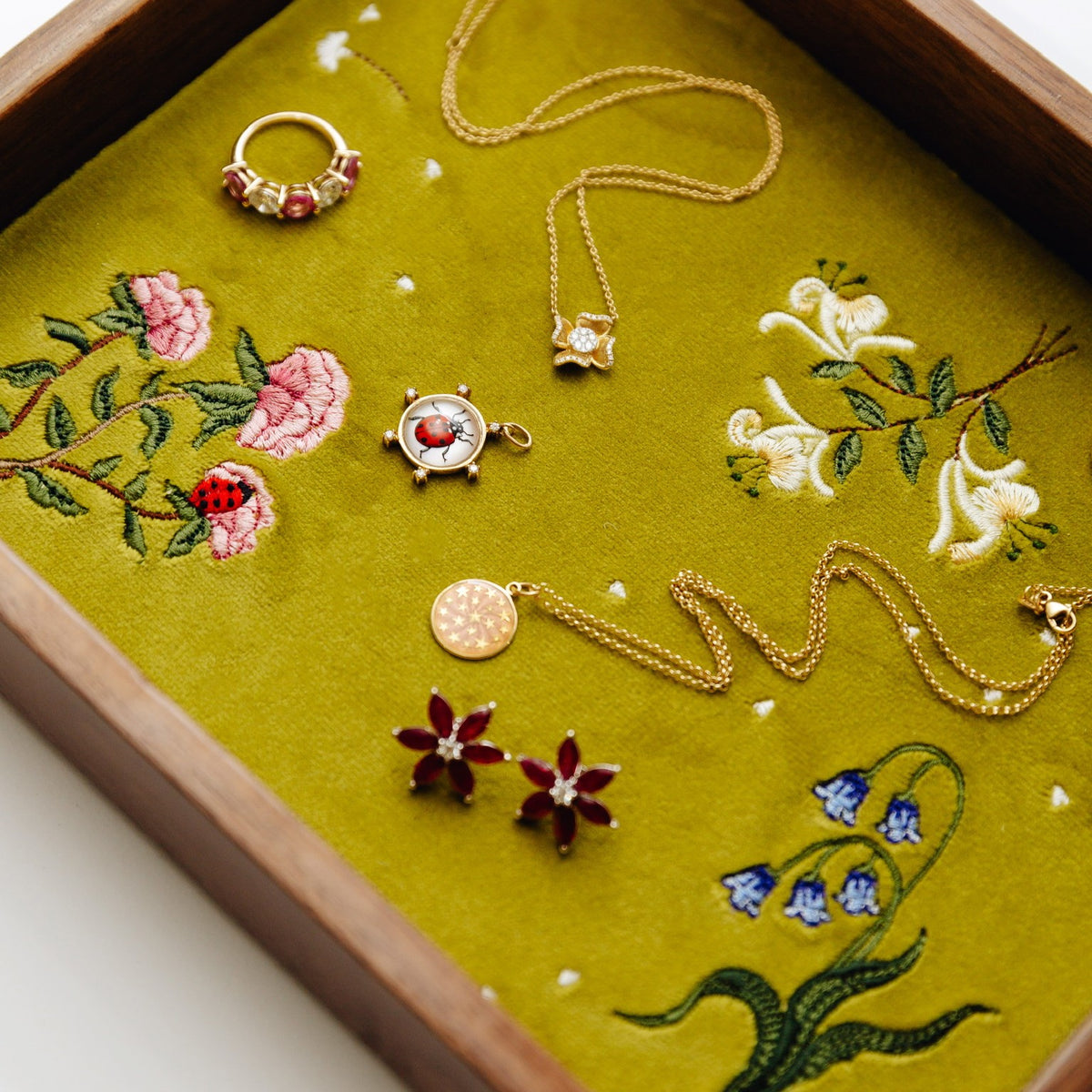 A styled flat-lay of assorted gold jewelry, including necklaces, earrings, and a ring, arranged on an olive green embroidered fabric insert inside a wooden jewelry tray.