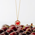 A gold chain necklace with a round pendant featuring a red heart on a light pink background, styled over a bed of dark red cherries against a white background.