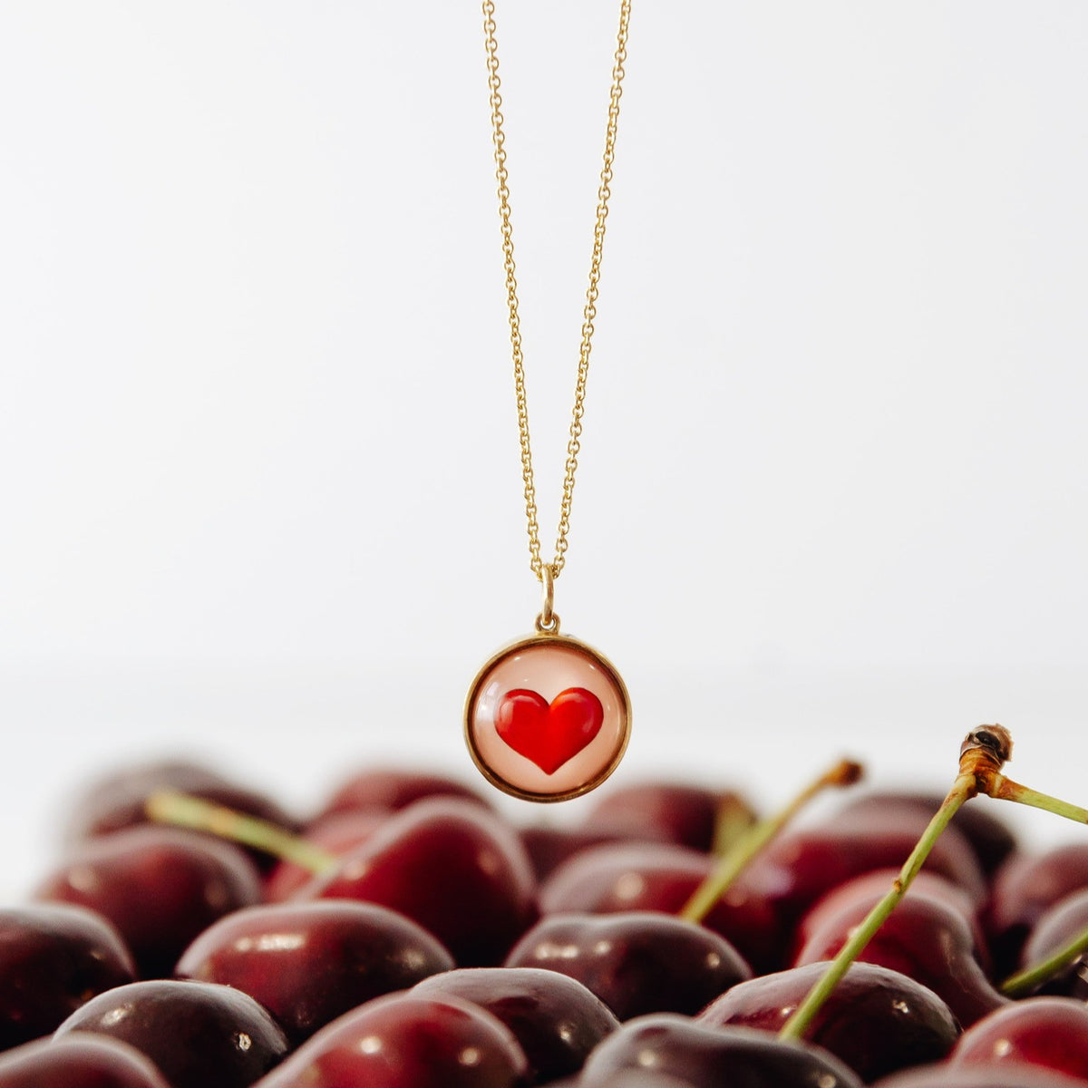 A gold chain necklace with a round pendant featuring a red heart on a light pink background, styled over a bed of dark red cherries against a white background.