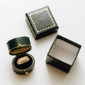 A dark green ring box set shown on a white background, including an open round box with a gold-trimmed velvet ring cushion holding a ring, a closed square box with gold decorative stitching, and an empty open square box.