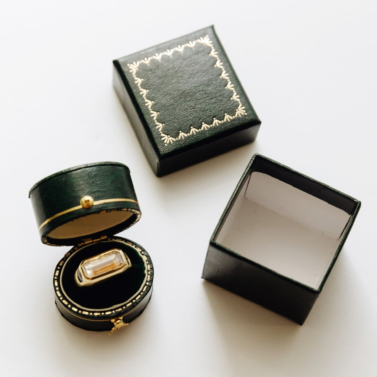 A dark green ring box set shown on a white background, including an open round box with a gold-trimmed velvet ring cushion holding a ring, a closed square box with gold decorative stitching, and an empty open square box.