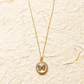 A gold chain necklace with a round pendant featuring a small orange and black butterfly on a white background, displayed on a textured beige paper backdrop.