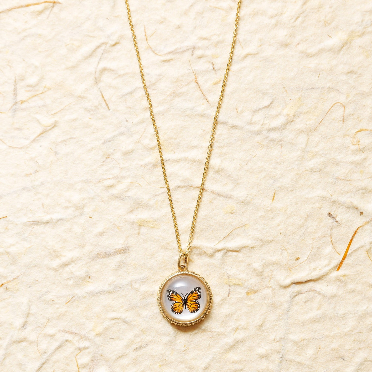 A gold chain necklace with a round pendant featuring a small orange and black butterfly on a white background, displayed on a textured beige paper backdrop.