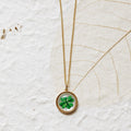 A gold chain necklace with a round pendant featuring a green four-leaf clover under glass, displayed on a textured white background with a dried leaf.