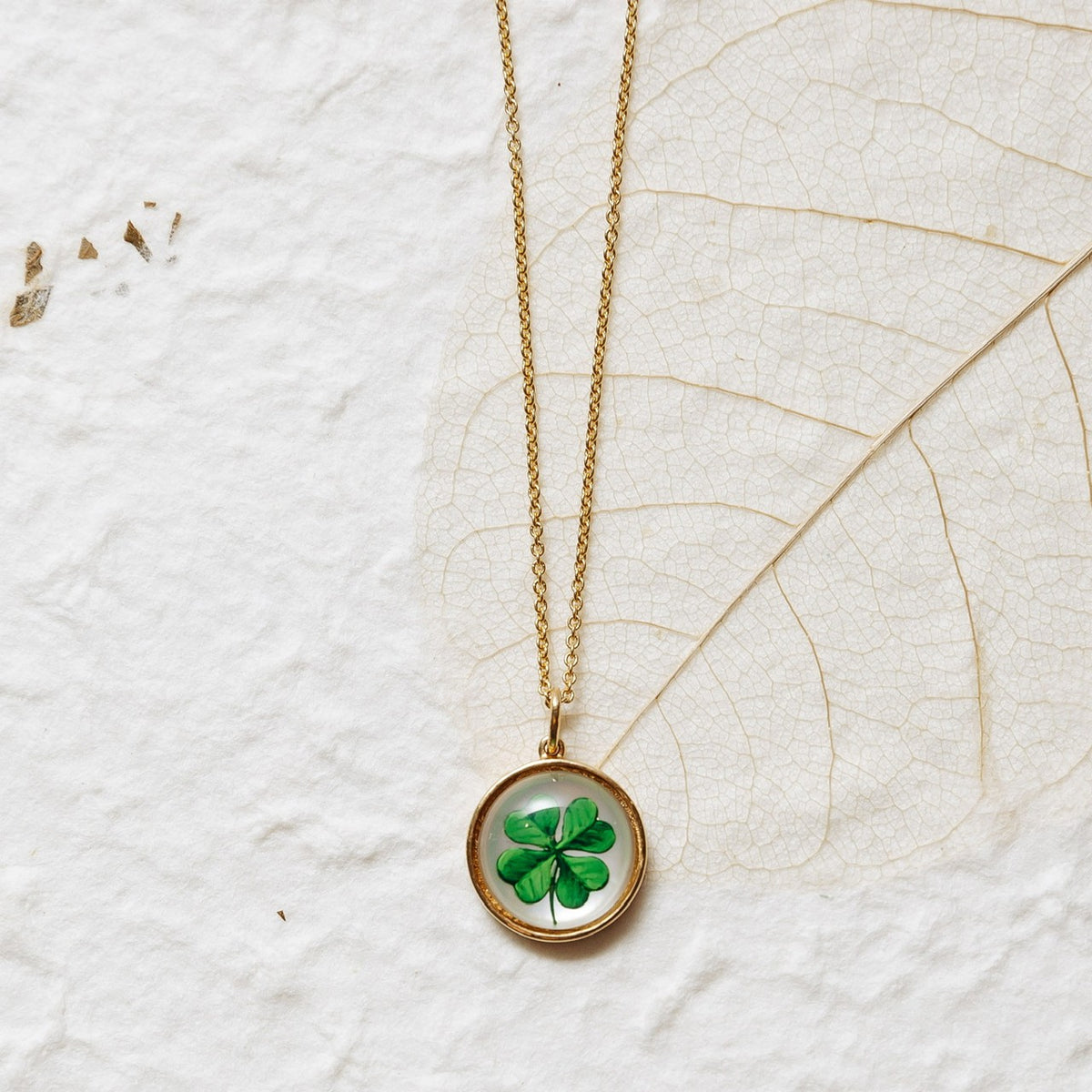 A gold chain necklace with a round pendant featuring a green four-leaf clover under glass, displayed on a textured white background with a dried leaf.