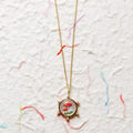 A gold chain necklace with a round pendant featuring two red-and-white spotted mushrooms on a textured off-white background with faint multicolored thread-like marks.