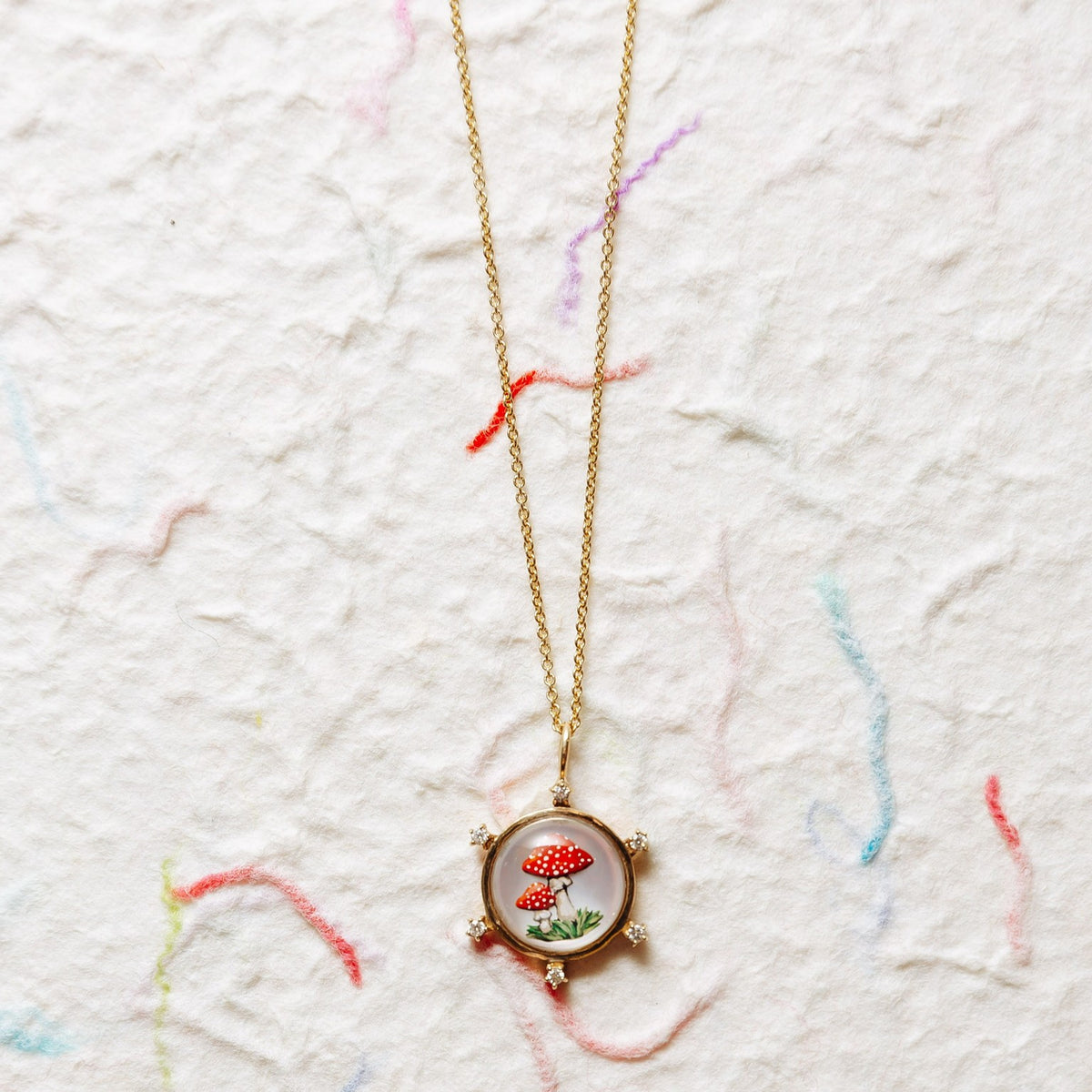 A gold chain necklace with a round pendant featuring two red-and-white spotted mushrooms on a textured off-white background with faint multicolored thread-like marks.