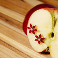 Two red flower-shaped stud earrings with clear center stones, displayed on a sliced red apple on a wooden surface.