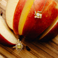 A gold flower-shaped pendant necklace and a matching gold sunburst ring with a yellow gemstone and clear stones, displayed on sliced red apples over a wooden surface.