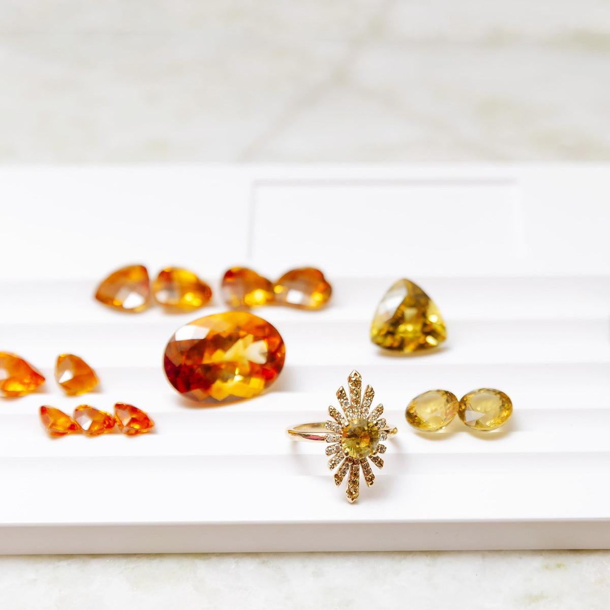 A collection of yellow and orange gemstone jewelry pieces, including a gold ring with a sunburst design arranged on a white display tray.