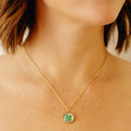 A close-up of a person's neck and upper chest wearing a delicate gold chain necklace with a round pendant featuring a green four-leaf clover on a white background.