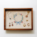A wooden jewelry tray with a beige velvet lining, featuring an embroidered floral wreath in the center and several pieces of jewelry arranged inside, including necklaces, earrings, and rings.