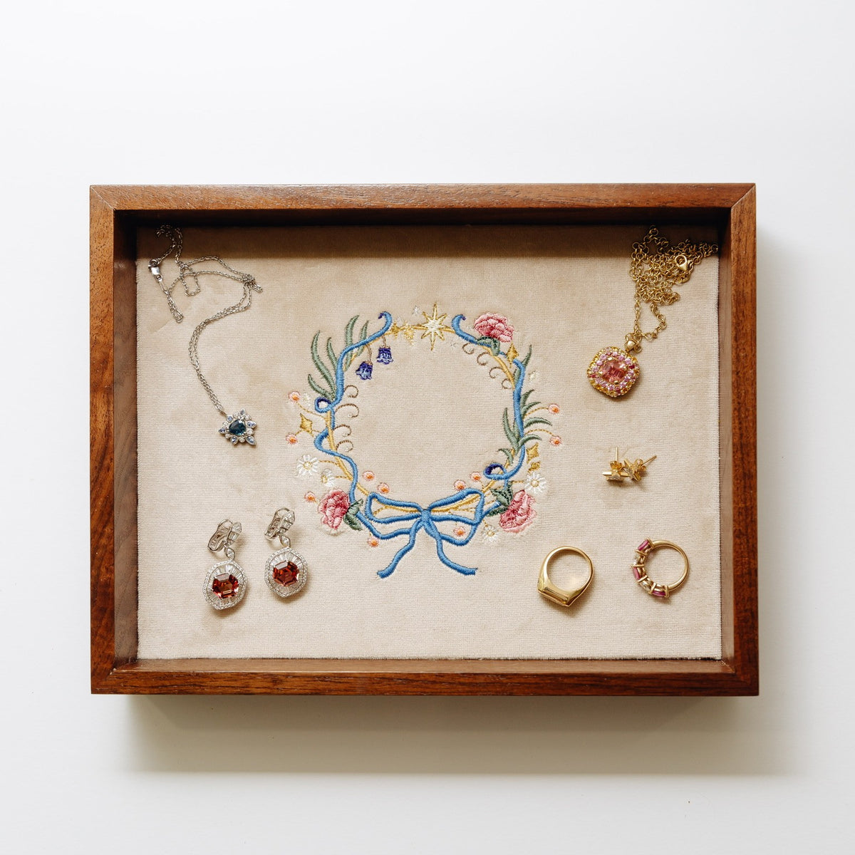 A wooden jewelry tray with a beige velvet lining, featuring an embroidered floral wreath in the center and several pieces of jewelry arranged inside, including necklaces, earrings, and rings.