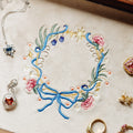 A close-up flat lay of a cream fabric surface featuring a colorful embroidered floral wreath with blue ribbon, pink flowers, green leaves, and gold star details. Surrounding the wreath are partial jewelry pieces, including a silver necklace, a peach gemstone pendant, a gold ring, and other earrings, arranged as styled props.