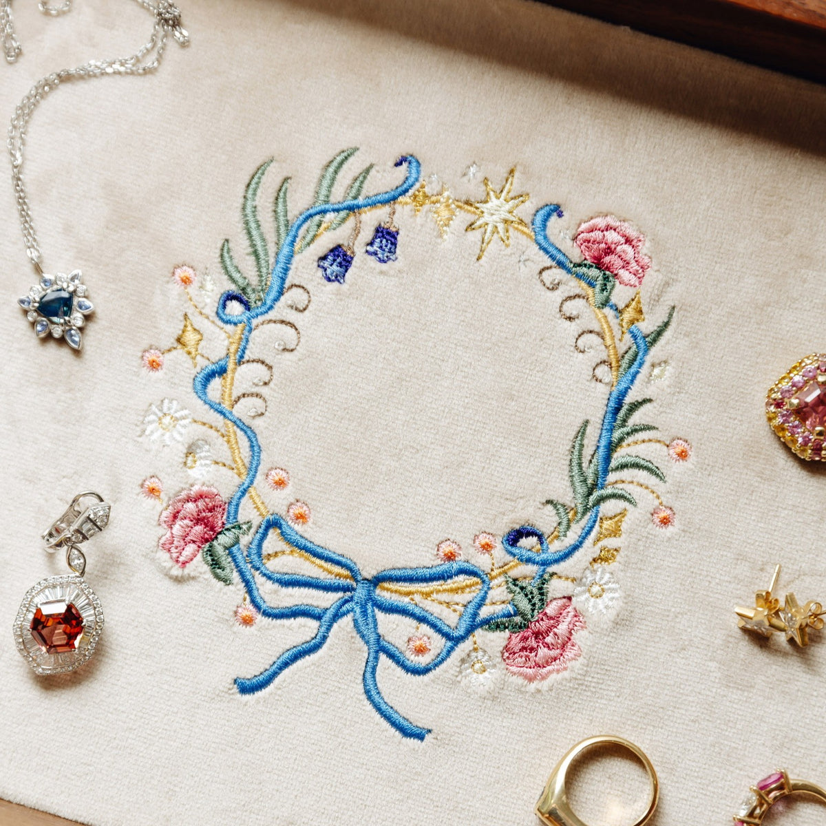 A close-up flat lay of a cream fabric surface featuring a colorful embroidered floral wreath with blue ribbon, pink flowers, green leaves, and gold star details. Surrounding the wreath are partial jewelry pieces, including a silver necklace, a peach gemstone pendant, a gold ring, and other earrings, arranged as styled props.