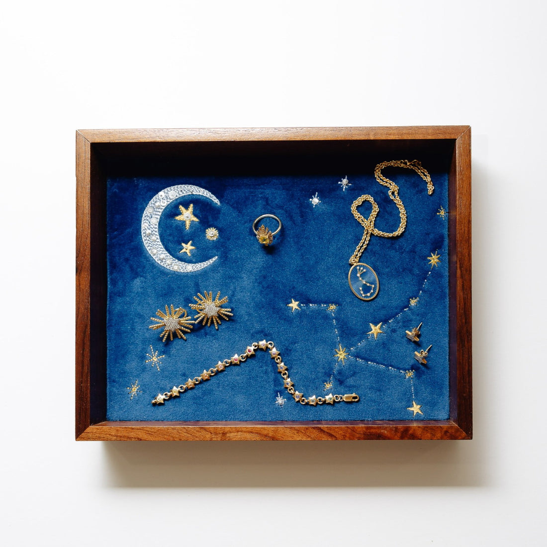 A square wooden jewelry tray containing a deep blue velvet background with embroidered celestial designs, including a crescent moon, stars, and constellations, set against a light beige background.