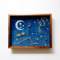 A jewelry tray display featuring celestial jewelry on a deep blue velvet background with embroidered stars and constellations, photographed against a plain white wall.