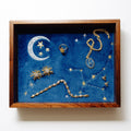 A square wooden shadow box display featuring celestial jewelry on a deep blue velvet background, mounted on a plain white wall.