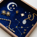 A curated collection of celestial-themed jewelry displayed inside a jewelry tray lined with deep blue velvet. The jewelry includes gold starburst stud earrings, a gold crescent moon and stars pendant, a gold ring with a pearl, a gold necklace with a silver-toned oval pendant, and a gold chain bracelet, all decorated with small star details.