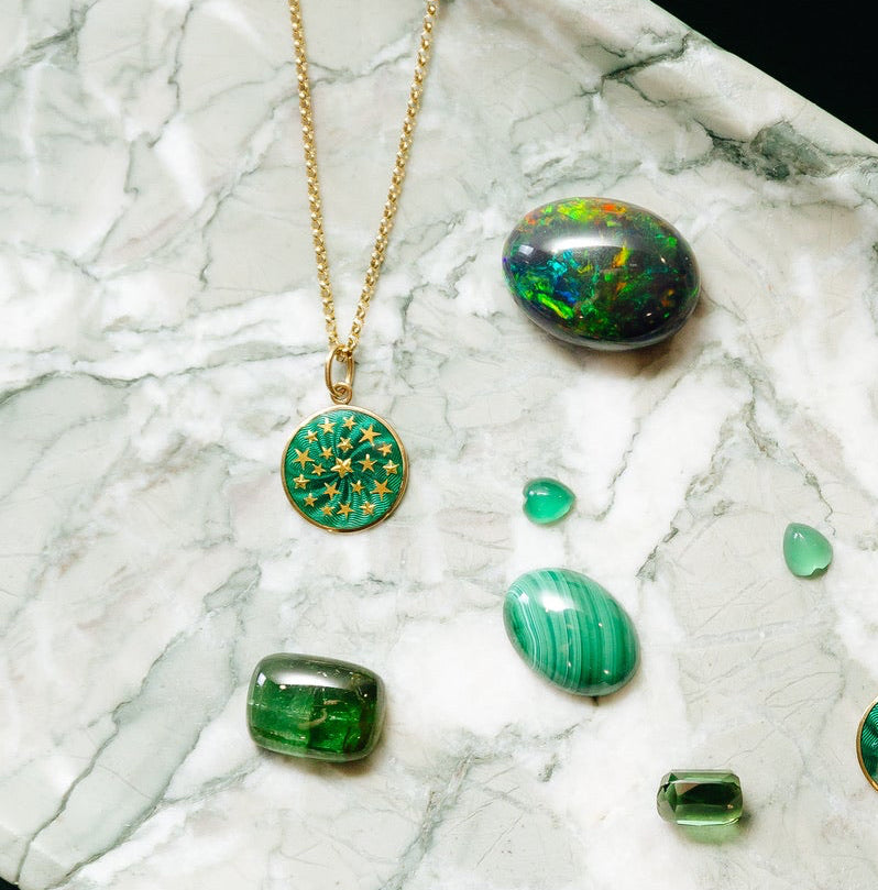 A gold necklace is displayed on a white marble surface. Its fine chain extends from the top edge of the image to a round pendant near the center-left. The pendant has a gold-toned rim and a glossy teal-green face decorated with numerous small gold stars in varying sizes. Several loose polished green gemstones surround the necklace, including a dark iridescent stone at the upper right and a striped green oval stone below it.