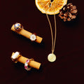 A flat-lay arrangement of jewelry and natural decorative elements on a dark burgundy background. Two bright orange dried citrus slices overlap near the upper center, with brown pinecones at the upper left and right. A gold beaded chain drapes from the citrus slices toward the lower center, ending in a round, pale-gold pendant with a spiral-like design. Two wooden cylinders lie diagonally across the left half of the image, each displaying assorted jewelry and rings.