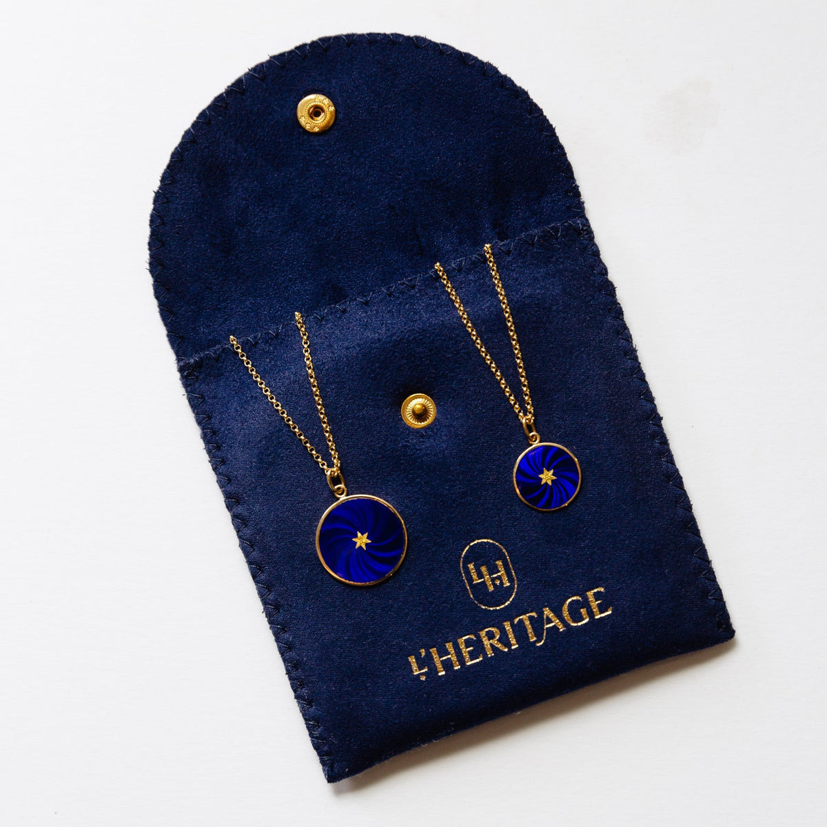 A dark navy fabric jewelry pouch lies diagonally on a white background. The pouch has a rounded flap, stitched edges, and a textured, suede-like surface. Two gold chains rest across the front, each attached to a circular blue pendant with a gold rim, a radiating swirl pattern, and a small gold star-shaped center. Gold snap fasteners are visible on the flap and lower section. Near the bottom is a gold oval monogram containing stylized letters, followed by the brand name “L’HERITAGE.”