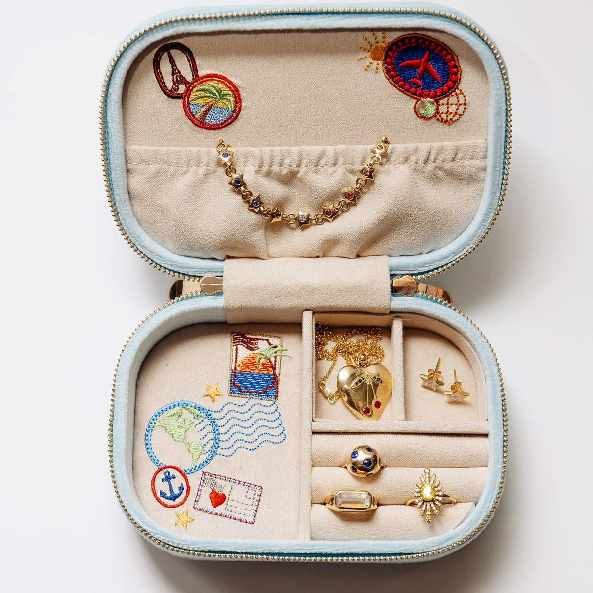 Open pale blue travel jewelry case displaying embroidered travel motifs and assorted gold-toned jewelry.