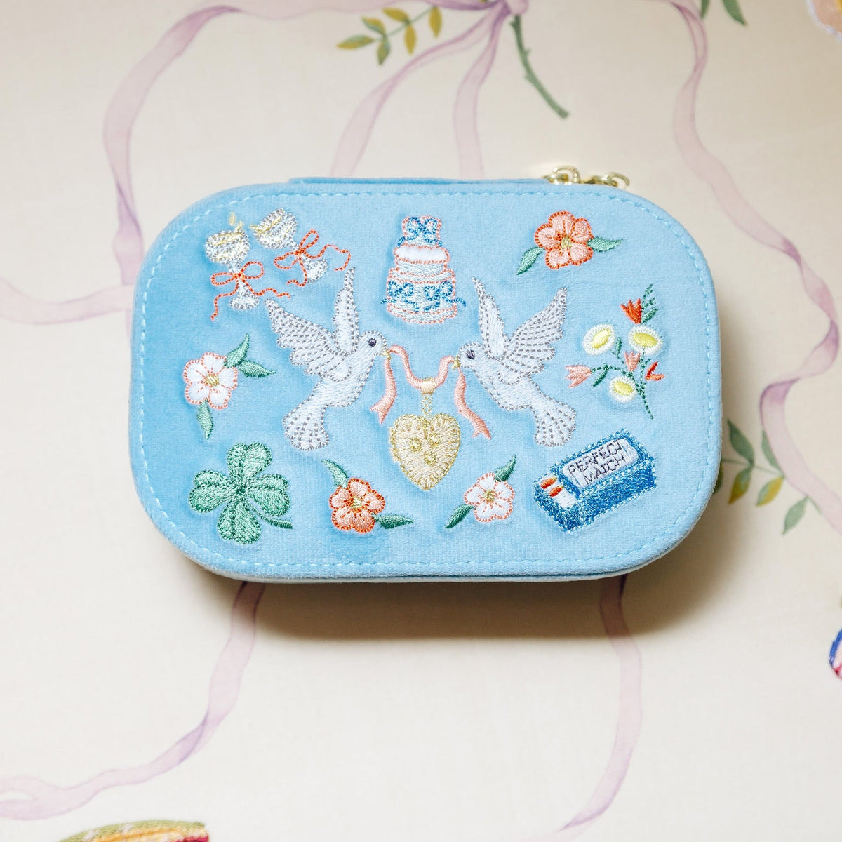 A small light-blue travel jewelry box with rounded corners and a zippered top rests on a pale floral-patterned surface. Its fabric exterior is decorated with colorful embroidery, including two white birds facing each other while holding a pink ribbon attached to a gold heart, small flowers and leaves, a four-leaf clover, crowns, a tiered cake, and a blue suitcase-like emblem. The box has visible stitching around its edges and two metallic zipper pulls at the top.