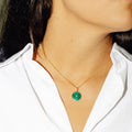 A close-up portrait shows the lower face, neck, and upper torso of a person with long dark hair. The person wears a white collared shirt and a delicate gold chain necklace with a round green pendant featuring a radiating spiral pattern and a small central accent. 