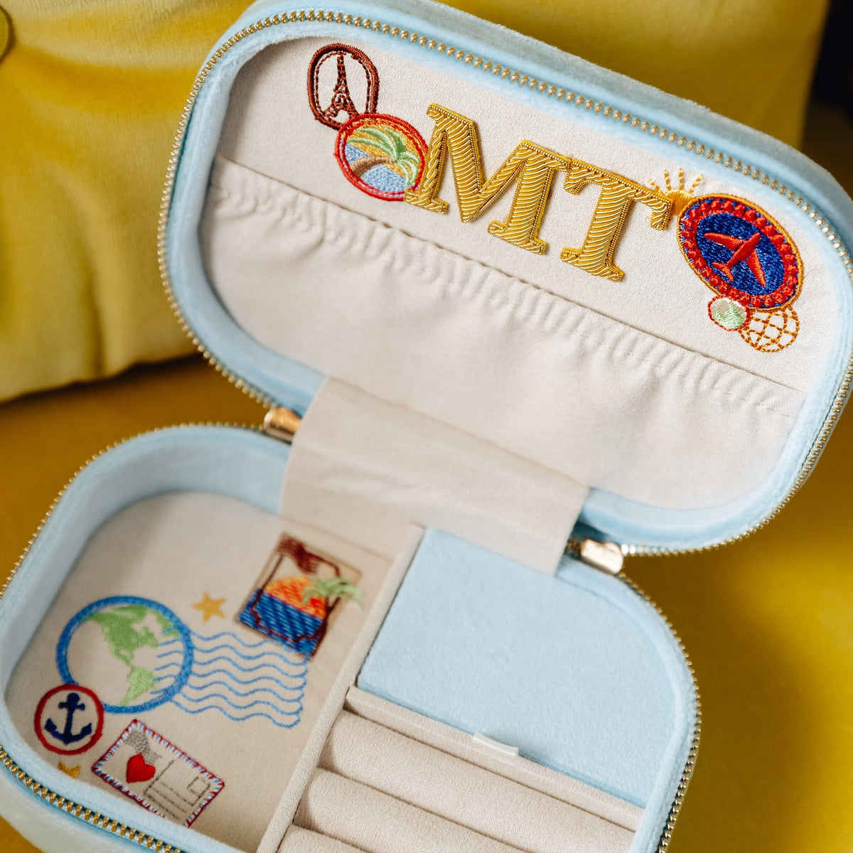 Open light-blue jewelry case with cream lining and colorful travel-themed embroidery, including a globe, anchor, airplane, and letters, "M," "T".

