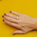 A close-up photograph centers on a gold-toned ring worn on a finger of a hand resting on a bright yellow surface. The ring has a slim band and a vertically oriented, elongated rectangular clear or white stone set in a raised, rounded-rectangle setting with reflective edges. The hand has dark burgundy nail polish. A pale yellow garment with a button is visible along the top edge of the image.