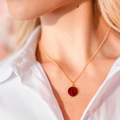 Person wearing a gold necklace with a red initial enamel guilloche pendant against a blurred background. Initial Guilloché Pendant. Crafted in 18k yellow gold with translucent guilloché enamel. 15mm diameter. Jewelry Dallas.