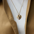 A gold heart-shaped pendant necklace with a small red cherry gemstone accent, shown on a model wearing a tan jacket over a white shirt.