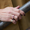 A close-up of a person's hand resting on a weathered metal railing, wearing a gold bracelet and a gold ring with pink sapphires and diamonds, set against a tan trench coat.