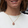 A woman with short brown hair wearing a white crewneck sweatshirt, and a gold pendant necklace with a pink stone, standing on a blurred city street.