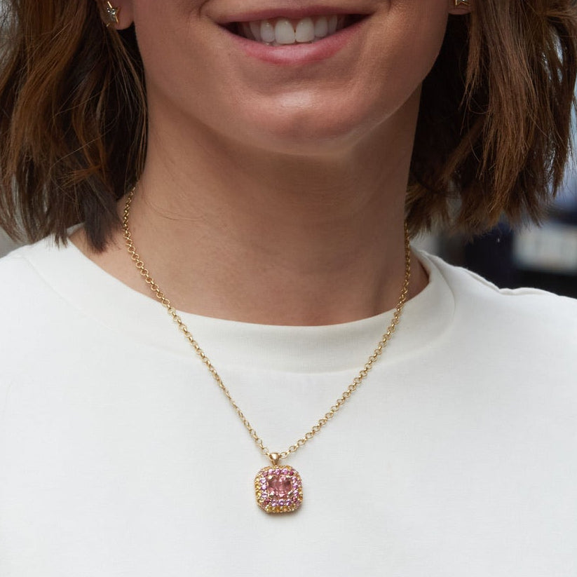A woman with short brown hair wearing a white crewneck sweatshirt, and a gold pendant necklace with a pink stone, standing on a blurred city street.