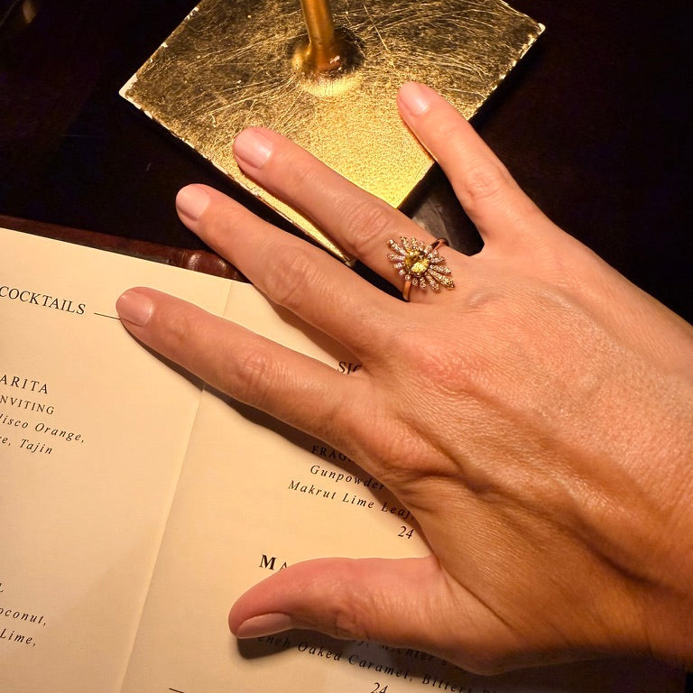 A close-up of a person's hand resting on an open cocktail menu, wearing a gold ring with a sunburst design and a central oval gemstone.