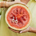 Watermelon with embedded earrings held by a person wearing a yellow dress. Pink Tourmaline Hexagon Drop Earrings with Diamonds. 9.10 carats of vibrant hexagon-cut pink tourmalines, accented by 2.84 carats of brilliant diamonds, set in 18k white gold. Jewelry Dallas.