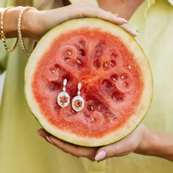 Watermelon with embedded earrings held by a person wearing a yellow dress. Pink Tourmaline Hexagon Drop Earrings with Diamonds. 9.10 carats of vibrant hexagon-cut pink tourmalines, accented by 2.84 carats of brilliant diamonds, set in 18k white gold. Jewelry Dallas.