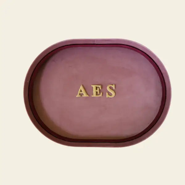 Oval-shaped pink velvet jewelry tray with 'AES' in gold on a beige background. Pink Velvet Jewelry Tray. Jewelry Dallas.