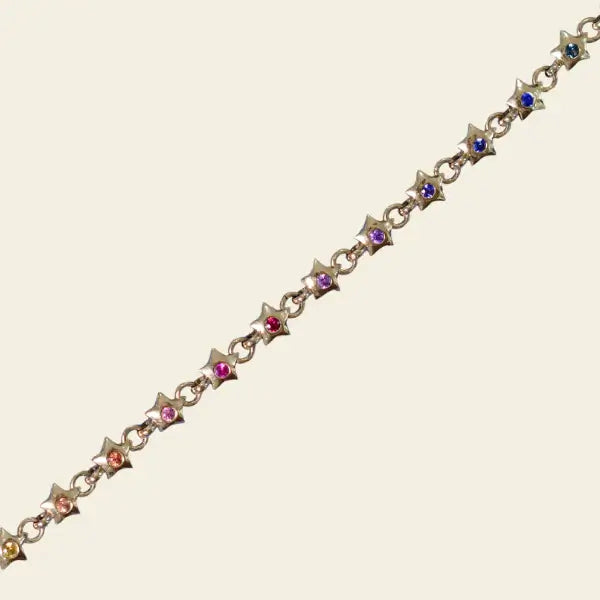 Gold bracelet with star-shaped charms and rainbow gemstones on a beige background. Rainbow Star Bracelet. Crafted in solid 14K yellow gold, each petite star is set with vibrant 0.80 carats of colored sapphires. Jewelry Dallas.