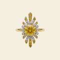 A yellow gold sunburst ring featuring a large round yellow center stone and smaller yellow and clear accent stones, set against a solid cream background.