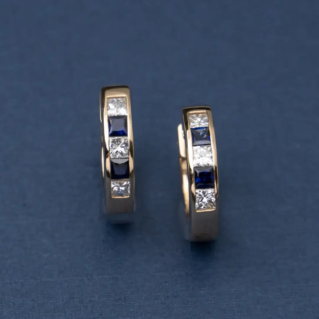 Gold huggie earrings with blue sapphires and diamonds on a beige background. Sapphire Checkered Huggies. Solid gold with natural sapphires and diamonds. Jewelry Dallas.