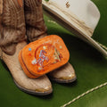 Cowboy boots with an orange embroidered Texas western travel jewelry box and a white hat on a green surface. Spirit of Texas Burnt Orange Velvet Embroidered Travel Jewelry Box. Jewelry Dallas.