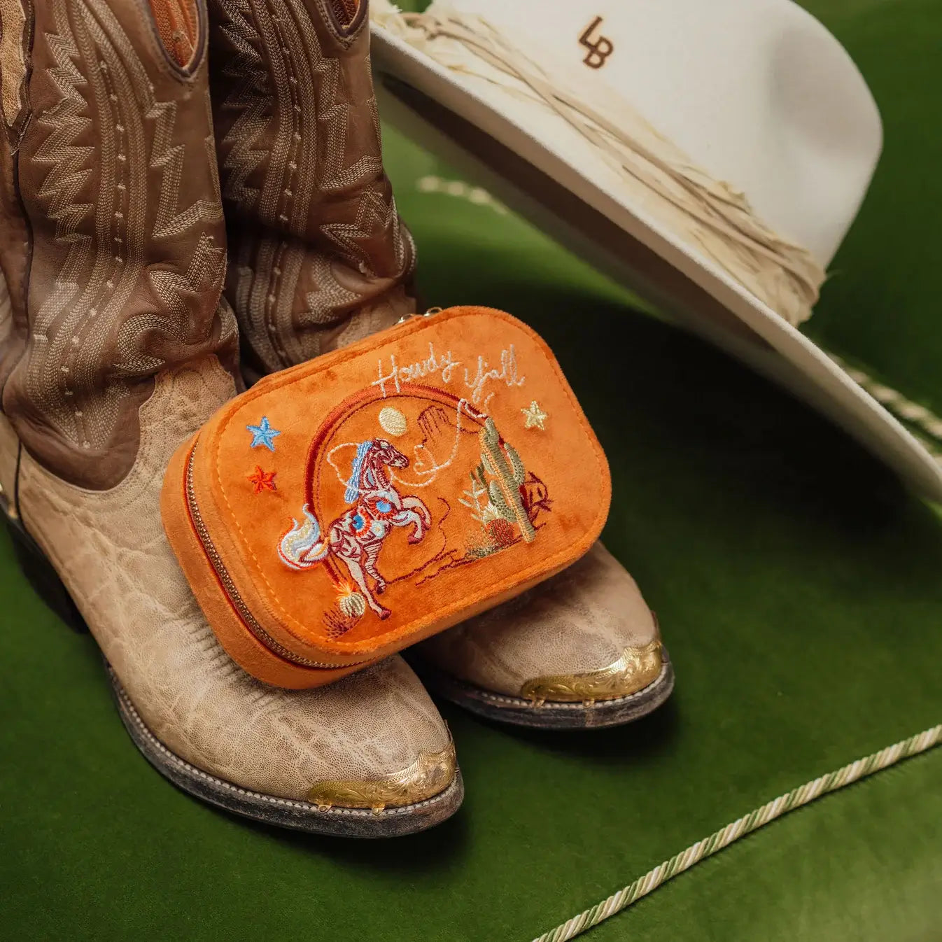 Cowboy boots with an orange embroidered Texas western travel jewelry box and a white hat on a green surface. Spirit of Texas Burnt Orange Velvet Embroidered Travel Jewelry Box. Jewelry Dallas.