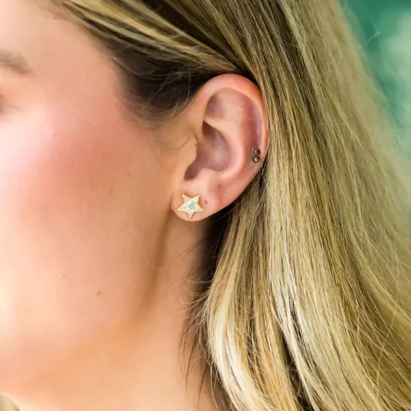 Close-up of an ear wearing a star-shaped gold and diamond earring with a blurred background. Star Studs. Materials: 18K Yellow Gold and Hand-Cut Natural Diamonds totaling 0.74 carats. Jewelry Dallas.
